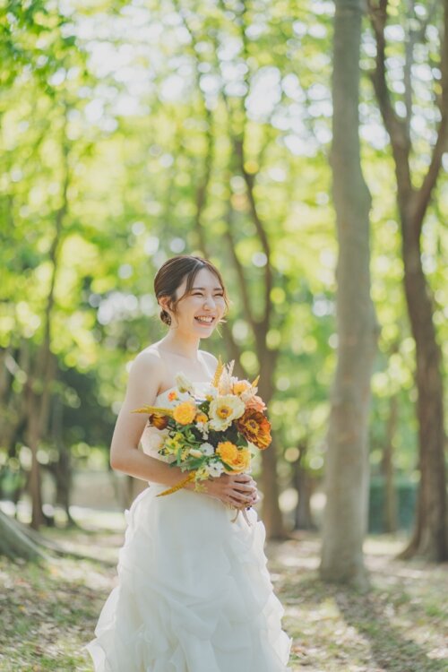 新緑の林でオレンジのブーケを胸元に抱え、笑顔を見せるフリルのウェディングドレスの花嫁。ロケーションフォトの一枚