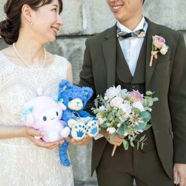 ぬいぐるみとブーケを持って笑い合うオリーブのスーツの新郎とレースのドレスの花嫁。洋装前撮りの小物