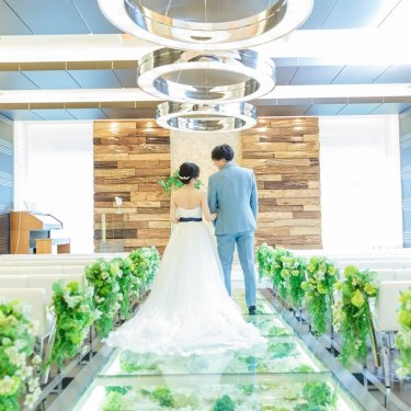 グリーンが並ぶガラスのバージンロードを腕を組んで歩く新郎新婦の後ろ姿。チャペルフォトの一枚