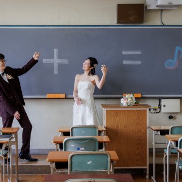 学校の教室で黒板の前に立ちポーズを決める新郎とウェディングドレスの花嫁。遊び心ある洋装前撮り