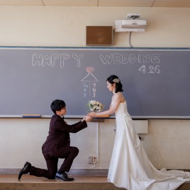 HAPPY WEDDINGと書いた黒板の前でひざまずいてブーケを渡す新郎。教室で撮るプロポーズの前撮り