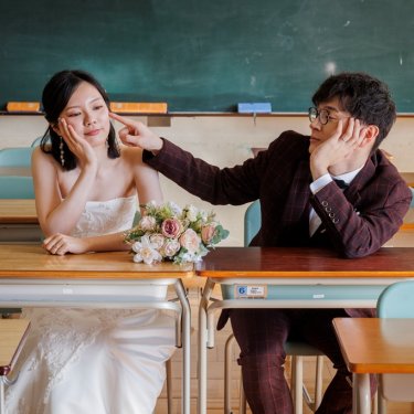 教室の机に並んで座り花嫁の頬をつつくチェックのスーツの新郎。学校で撮る洋装ロケーションフォト