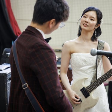 マイクスタンドの前でギターを持つウェディングドレスの花嫁と新郎。学校の音楽室で撮る遊び心ある前撮り