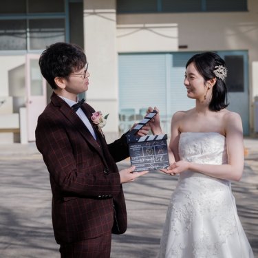 学校の校庭でカチンコを持って向き合うチェックのスーツの新郎とウェディングドレスの花嫁の前撮り
