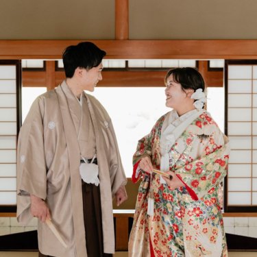 障子の座敷で扇子を手に顔を見合わせて笑う、ベージュの紋付袴の新郎と花柄の色打掛の花嫁の和装前撮り