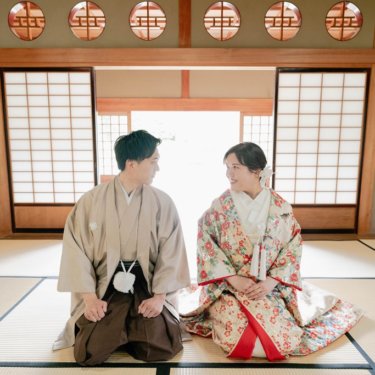 丸い欄間のある座敷の畳に並んで座り、顔を見合わせる紋付袴の新郎と花柄の色打掛の花嫁。日本家屋で撮る和装前撮り