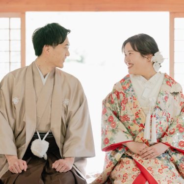 障子の明るい座敷に並んで座り、顔を見合わせて笑うベージュの紋付袴の新郎と花柄の色打掛の花嫁。大仙公園で撮る和装前撮り