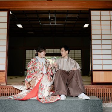 障子を開け放った日本家屋の縁に並んで腰かけ、笑い合う色打掛の花嫁とベージュの紋付袴の新郎の和装ロケーションフォト