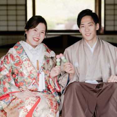 障子の座敷に並んで座り、三色団子を手に笑顔を見せる花柄の色打掛の花嫁とベージュの紋付袴の新郎の和装前撮り
