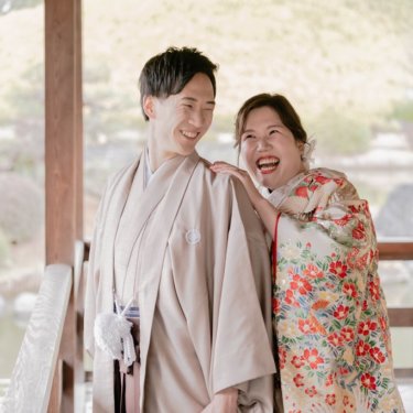 東屋で新郎の肩に手を添えて大笑いする花柄の色打掛の花嫁と、照れ笑いする紋付袴の新郎の和装ロケーションフォト