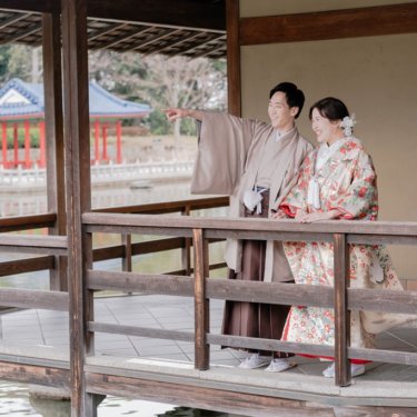 池に張り出した東屋の手すりのそばで、遠くを指さして笑う紋付袴の新郎と色打掛の花嫁。日本庭園の和装ロケーションフォト