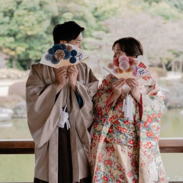 池を背にした東屋で扇子を広げて顔を隠す紋付袴の新郎と色打掛の花嫁、大仙公園で撮るおちゃめな和装前撮り
