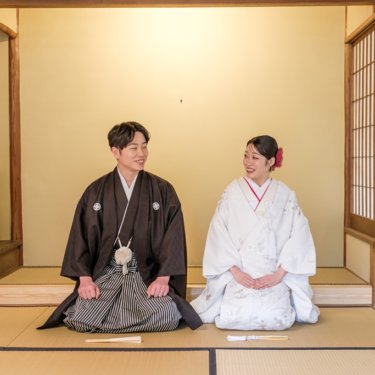 障子と床の間のある和室の畳に並んで座り、ほほえむ紋付袴の新郎と白無垢の花嫁の和装ロケーションフォト