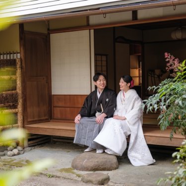 緑を前ぼかしに、日本家屋の縁側に並んで腰かける紋付袴の新郎と白無垢の花嫁。慶沢園の和装ロケーションフォト