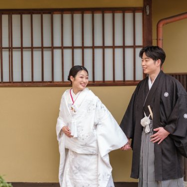 格子窓の日本家屋の前で手をつなぎ、顔を見合わせて笑う白無垢の花嫁と紋付袴の新郎の和装前撮り
