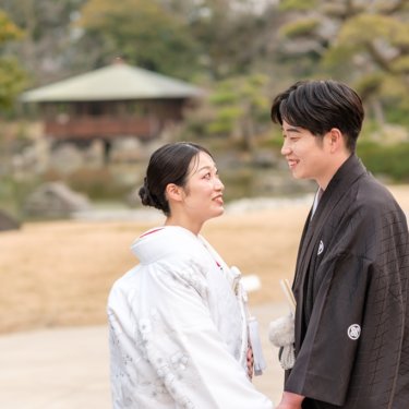 東屋が見える慶沢園で向き合い、ほほえみ合う紋付の新郎と白無垢の花嫁の和装ロケーションフォト