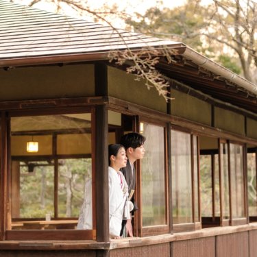 池に面した数寄屋造りの建物の窓辺に並び、外を眺める白無垢の花嫁と紋付の新郎。慶沢園の和装前撮り