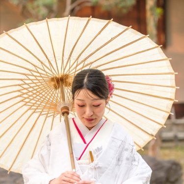 白い和傘を肩にかけ、手元を見つめてほほえむ白無垢の花嫁のソロカット。慶沢園で撮る和装前撮り