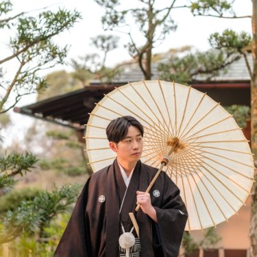 松と日本家屋を背に白い和傘を肩にかけて立つ紋付袴の新郎のソロカット、和装ロケーションフォト