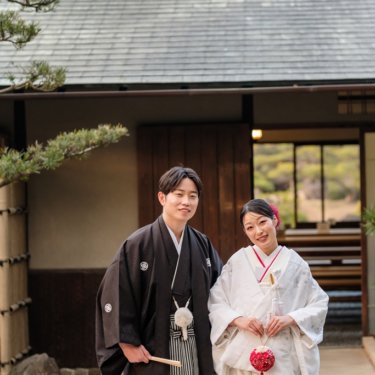 慶沢園にて、瓦屋根の日本家屋の前に並んで立ち、ほほえむ紋付袴の新郎と白無垢の花嫁の和装ロケーションフォト