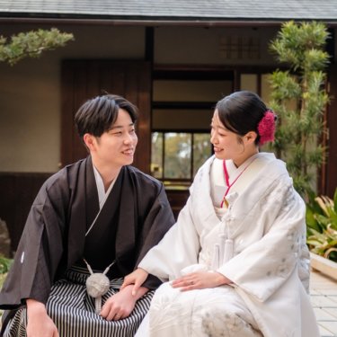 日本家屋の前に並んで座り、見つめ合う紋付袴の新郎と白無垢の花嫁。慶沢園で撮る和装ロケーションフォト