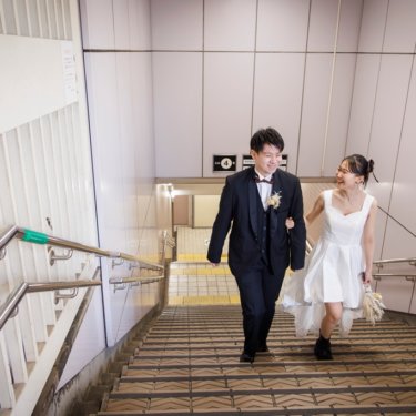 駅の階段を手をつないで駆け上がる黒のスーツの新郎とショート丈のドレスの花嫁、躍動感のある街ロケ前撮り