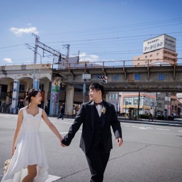 野田駅の高架を背に、青空の下の横断歩道を手をつないで渡る新郎とショート丈のウェディングドレスの花嫁の前撮り