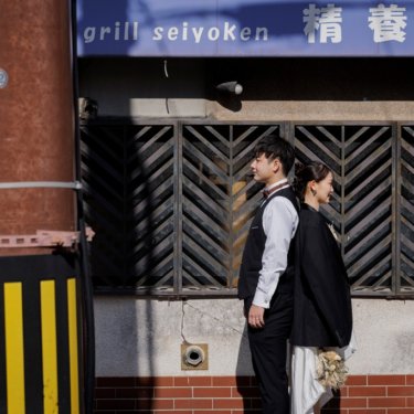 レトロな洋食店の格子窓の前で背中合わせに立つベスト姿の新郎とウェディングドレスの花嫁、街ロケの前撮り