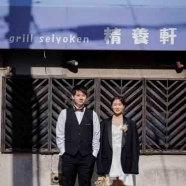 レトロな洋食店の格子窓の前に並んで立つベスト姿の新郎とジャケットを羽織ったショート丈のドレスの花嫁の前撮り