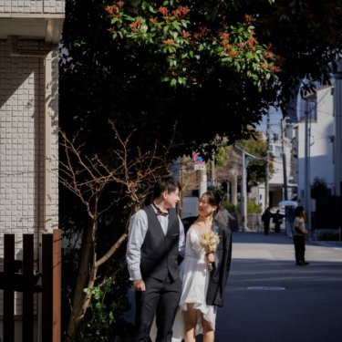木漏れ日の住宅街の歩道で腕を組んで歩くベスト姿の新郎とショート丈のウェディングドレスの花嫁の街ロケ前撮り