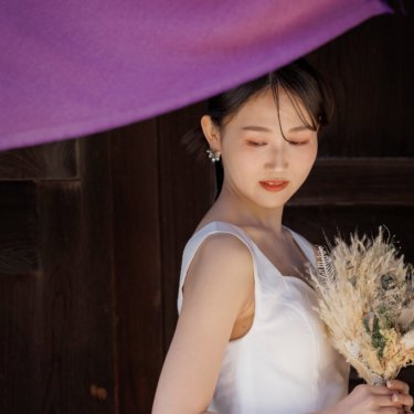 紫の幕の下でドライフラワーのブーケを持ち、目を伏せるショート丈のウェディングドレスの花嫁。神社の門で撮る前撮り
