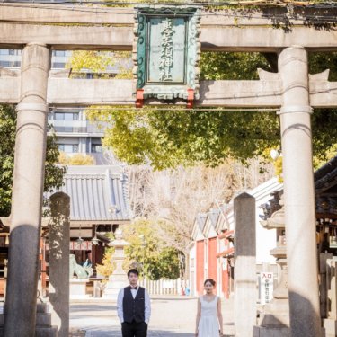 石の鳥居をくぐった神社の参道に、離れて立つベスト姿の新郎とショート丈のウェディングドレスの花嫁の前撮り