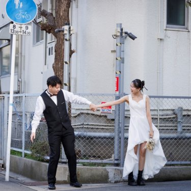 住宅街のフェンス沿いで手をつなぎ、引っ張り合って笑うベスト姿の新郎とショート丈のドレスの花嫁の街ロケ前撮り