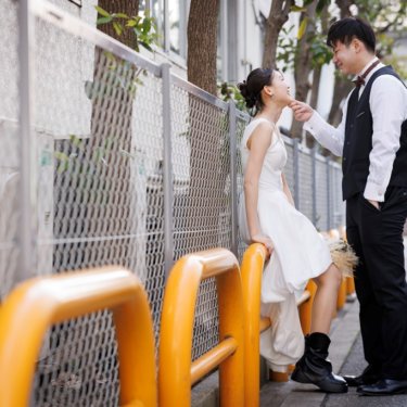 オレンジの柵に腰かける花嫁のあごに手を添え、見つめ合うベスト姿の新郎。街角で撮るロマンチックな前撮り