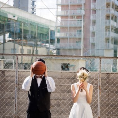フェンスの前でバスケットボールとブーケで顔を隠しておどける新郎とショート丈のドレスの花嫁、遊び心のある前撮り