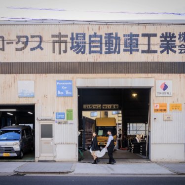 レトロな看板の自動車工場の入口で、手をつないで立つ新郎とショート丈のウェディングドレスの花嫁の個性派ロケーションフォト
