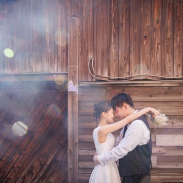 光が差し込む木の壁の前でおでこを寄せて抱き合う新郎と花嫁、レトロな路地で撮るロマンチックな前撮り