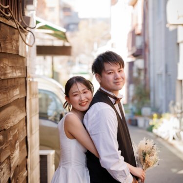 木の壁の路地で新郎に後ろから抱きつき、肩越しにほほえむウェディングドレスの花嫁。下町で撮る街ロケ前撮り
