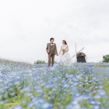 ネモフィラの花畑の先、風車を背に手をつないで立つ新郎とウェディングドレスの花嫁。鶴見緑地公園のロケーションフォト