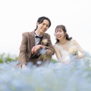 青いネモフィラの花畑にしゃがみ、ブーケを持って笑い合うツイードの新郎とウェディングドレスの花嫁の春の前撮り