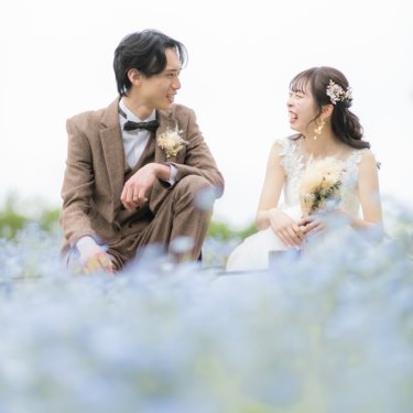 ネモフィラの花を前ぼかしに、しゃがんで顔を見合わせ笑う新郎とウェディングドレスの花嫁。鶴見緑地公園の前撮り