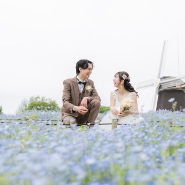 鶴見緑地にて、風車を背にネモフィラの花畑の中に座り、笑顔を交わすツイードの新郎とウェディングドレスの花嫁の洋装前撮り