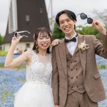 鶴見緑地にて、風車とネモフィラを背にハート形サングラスを掲げておどけるツイードの新郎とウェディングドレスの花嫁の前撮り