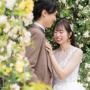 白いモッコウバラの前で新郎の胸に手を添え、顔を寄せて笑うウェディングドレスの花嫁。春の鶴見緑地公園の前撮り