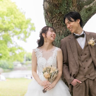 大木の幹にもたれ、顔を見合わせて笑うツイードの新郎とドライフラワーのブーケを持つ花嫁の洋装前撮り