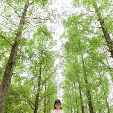 そびえるメタセコイア並木の下で、ブーケを持ってうつむくウェディングドレスの花嫁のソロカット。鶴見緑地公園の前撮り
