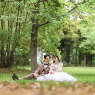 新緑の木立の芝生に並んで座り、くつろいで笑い合う新郎とウェディングドレスの花嫁を広く捉えた前撮り