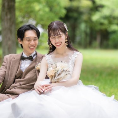 芝生にドレスを広げて並んで座り、手元を見てほほえむツイードの新郎とウェディングドレスの花嫁の前撮り