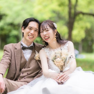 鶴見緑地にて、新緑の公園の芝生に座り、ほおを寄せて満面の笑みを見せるツイードの新郎とウェディングドレスの花嫁の前撮り