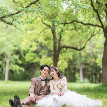 落ち葉を前ぼかしに、新緑の木の下の芝生に並んで座る新郎とウェディングドレスの花嫁。春の公園の洋装前撮り